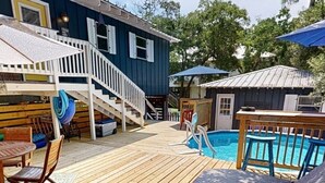 A wooden deck with a pool, outdoor furniture, and a staircase leading to a blue building. - Wooded area home with private pool, pool table, and outdoor amenities (Dauphin Island)