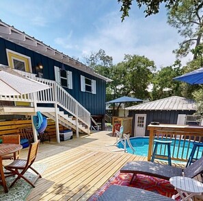 Eine Holzterrasse mit Pool, Gartenmöbeln und einer Treppe zu einem blauen Gebäude.