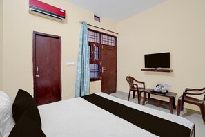 A hotel room with a bed, a desk with a chair, a television, and an air conditioning unit. - Classic Ganesha (Kharar)