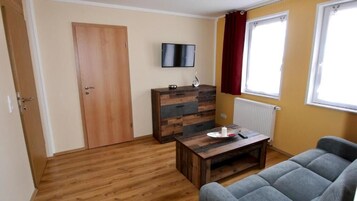 Una habitación con suelo de madera, un sofá, un aparador de madera, un televisor en la pared y una ventana con cortinas.