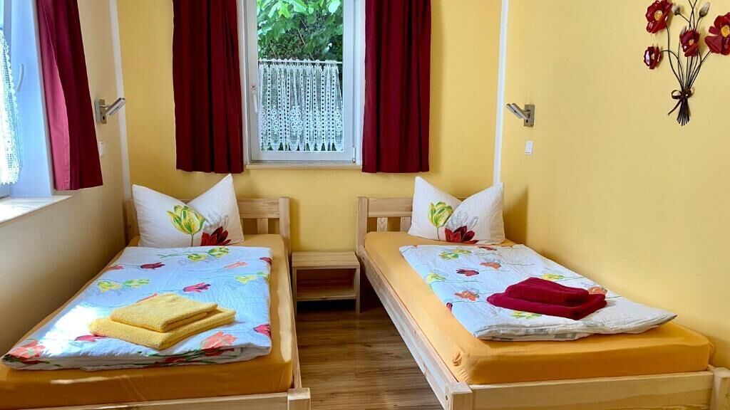 Une chambre avec deux lits en bois, chacun agrémenté d’une couvre-lit et de coussins à motifs floraux, une fenêtre avec des rideaux rouges et une décoration murale représentant des fleurs rouges.