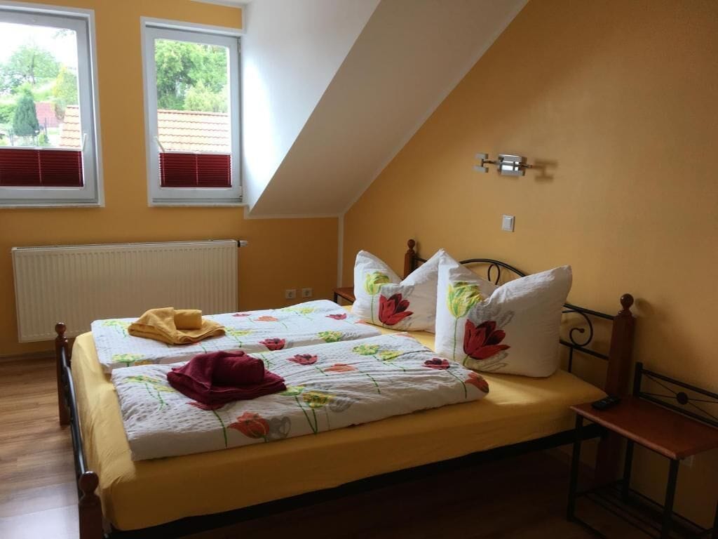A bedroom with a sloped ceiling, a bed with floral bedding, two windows with shutters, and a radiator.