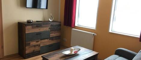 Una habitación con una televisión de pantalla plana montada en la pared, un aparador de madera con cajones, un sofá gris, una mesita con una vela y flores, y una ventana con cortinas rojas.