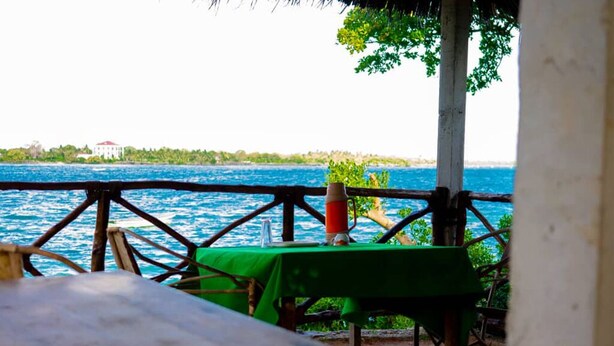 A table with a green tablecloth, a red bottle, and a glass on a wooden table, set against a backdrop of a blue sea and a white building in the distance.