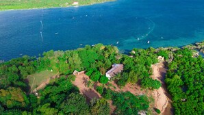 An aerial view of a coastal area with dense greenery, a small building, and a clear blue sea with boats.