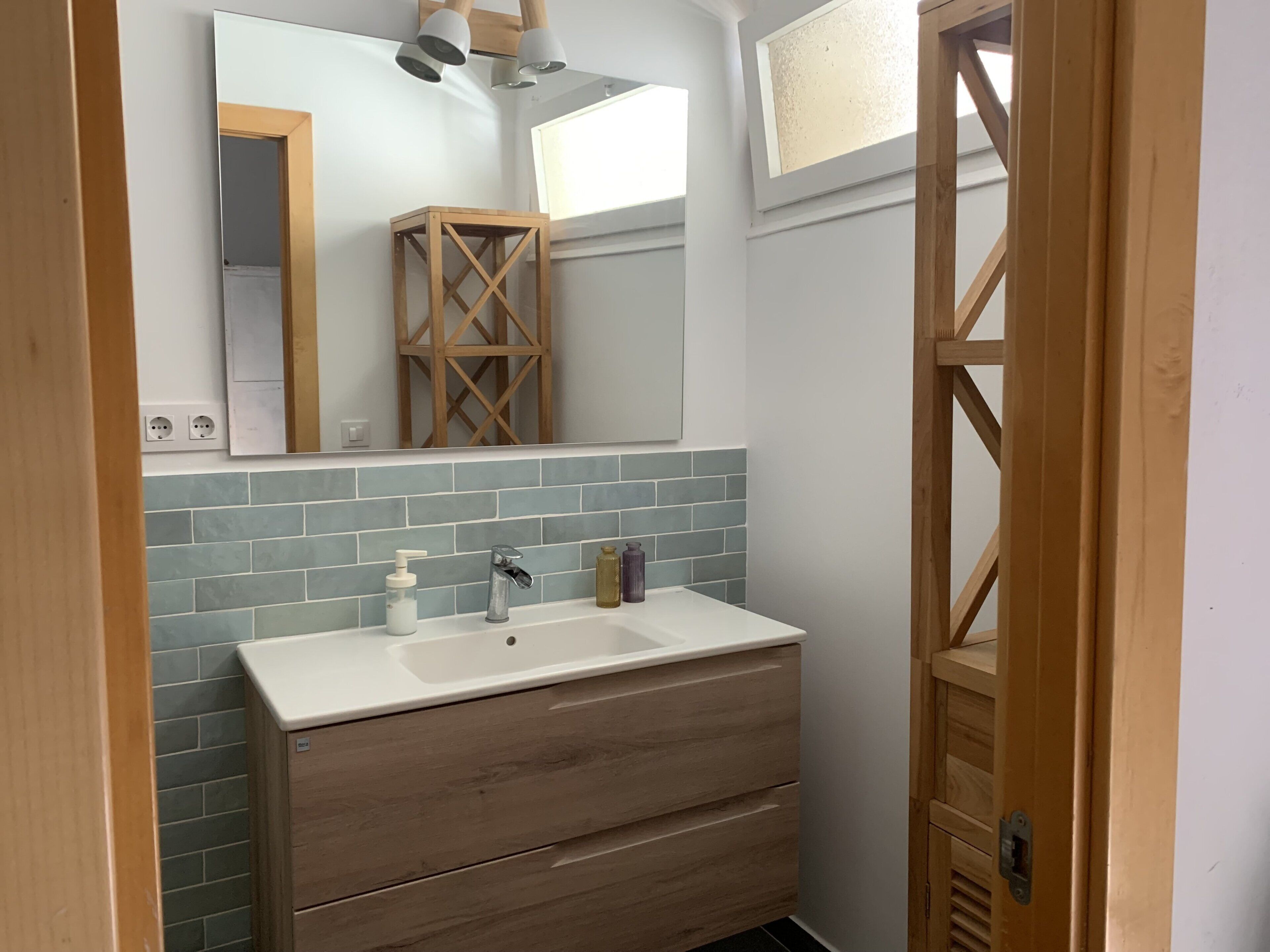 Un baño con lavamanos blanco, tocador de madera, salpicadero con azulejos azules y un espejo sobre el lavamanos.