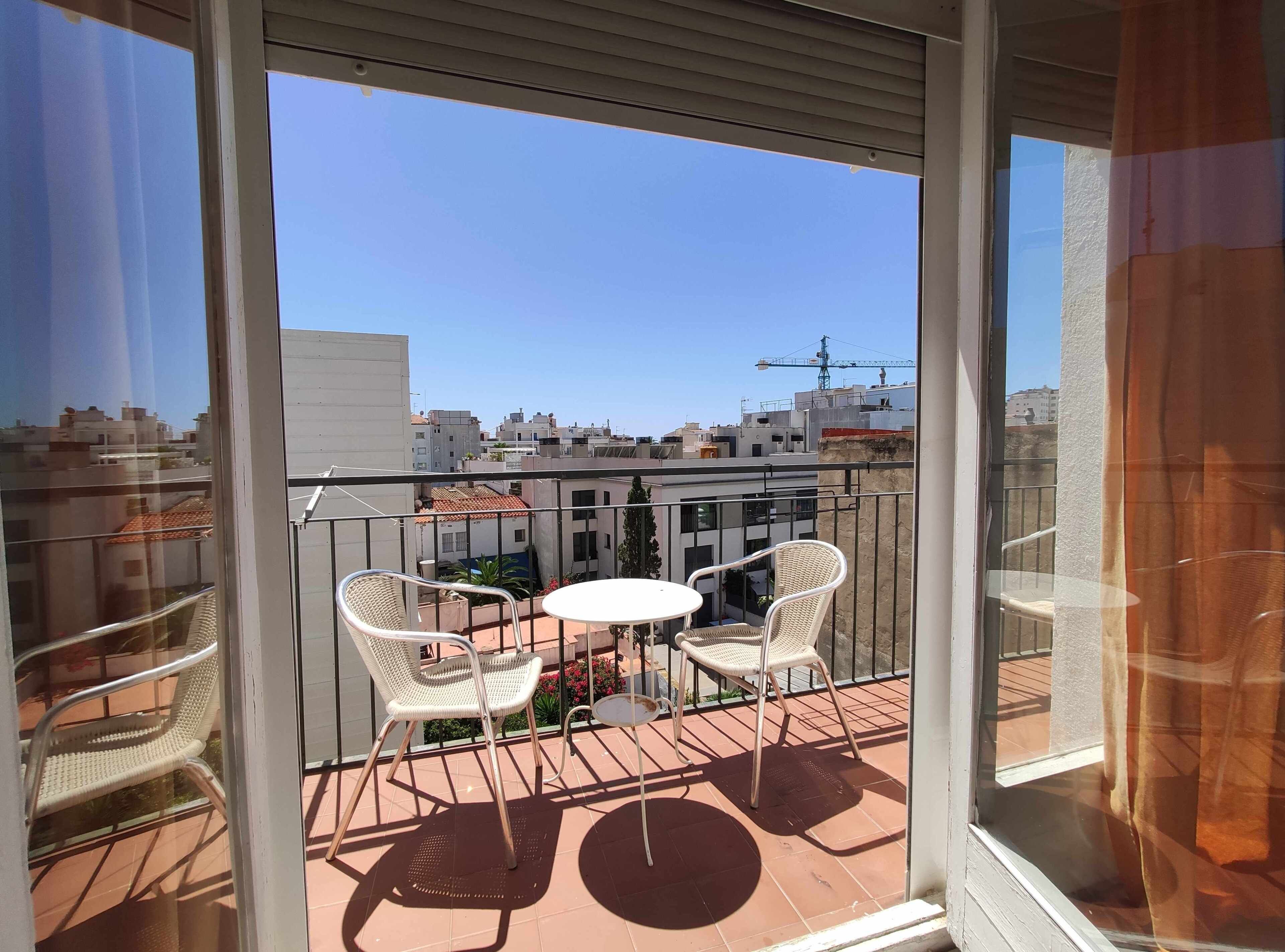 Un balcón con dos sillones de mimbre y una pequeña mesa redonda, con vistas a un paisaje urbano que incluye edificios y una grúa de construcción.