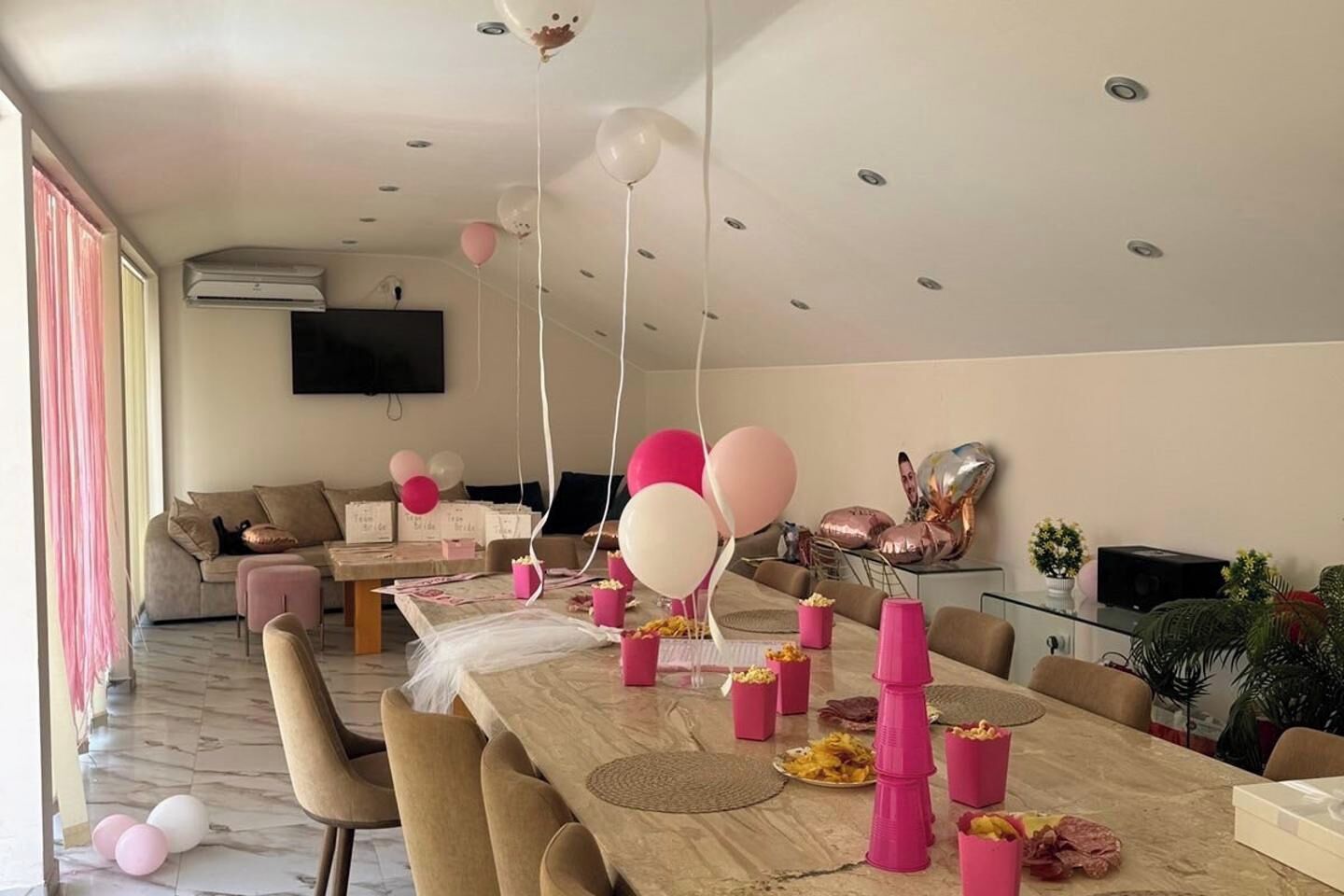 Una sala moderna con una larga mesa de comedor, globos rosa y blancos y un televisor de pantalla plana montado en la pared.