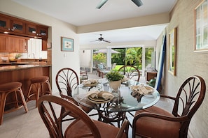 A dining area with a glass table surrounded by wicker chairs, a kitchen with wooden cabinets, and a view of a garden through large windows. - Garden Condo Near Poipu Beach | Pool, Hot Tub & Tennis | Manualoha 203 (Koloa)