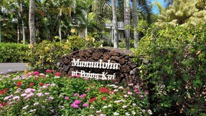 A sign with "Manu'oloha at Poipu Kai" written on it, surrounded by lush greenery and flowering plants. - Garden Condo Near Poipu Beach | Pool, Hot Tub & Tennis | Manualoha 203 (Koloa)