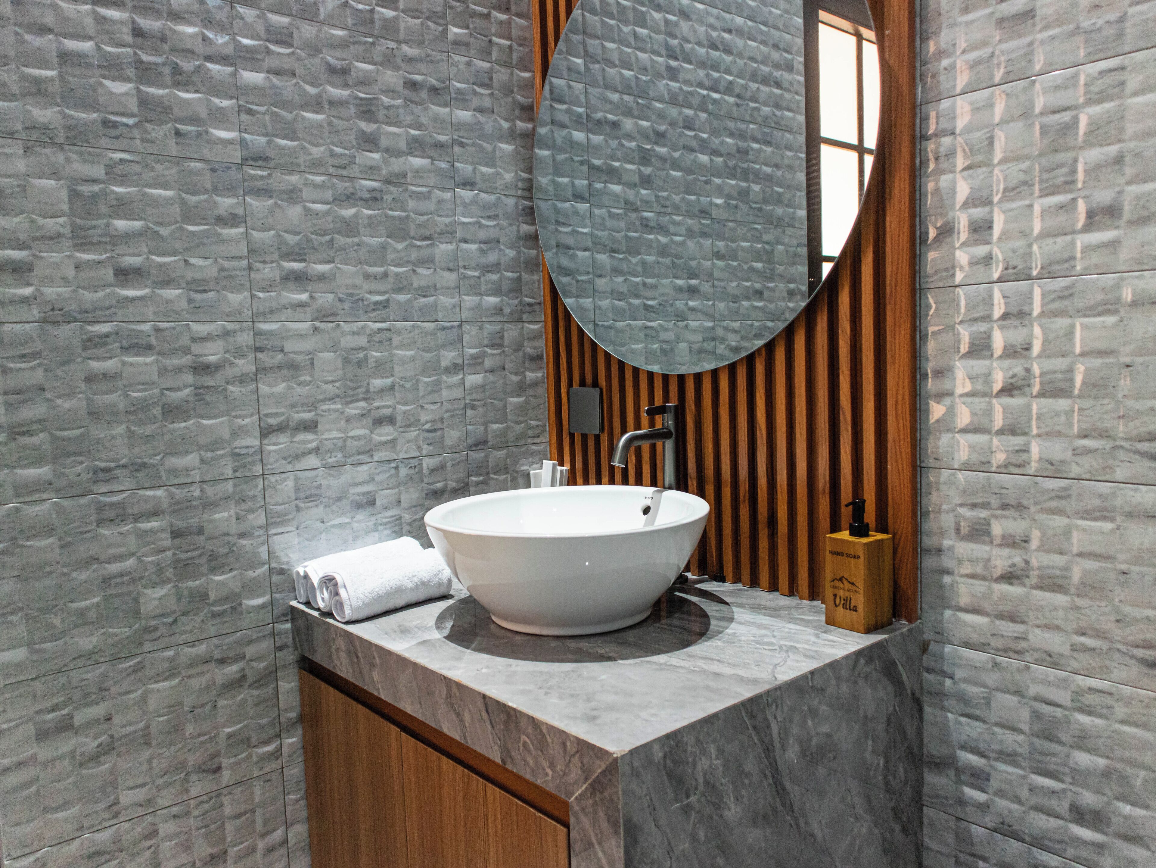 A bathroom with a round mirror, a white sink, wooden paneling, and a marble countertop.