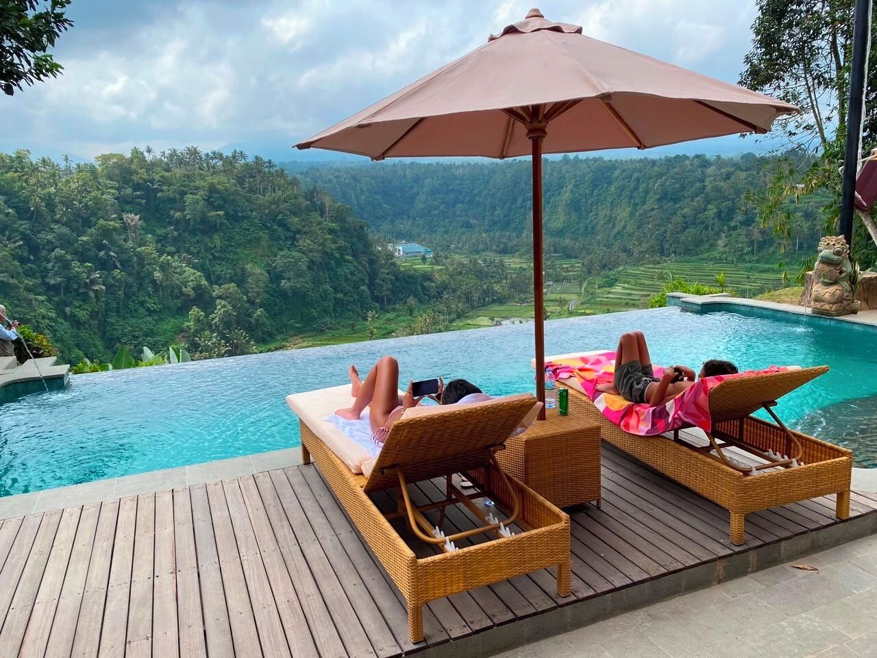 A wooden deck with wicker lounge chairs and a large umbrella overlooking a swimming pool with a view of a lush landscape.
