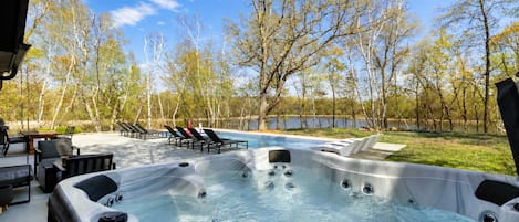 A hot tub area with a pool and lounge chairs, surrounded by trees and a body of water.
