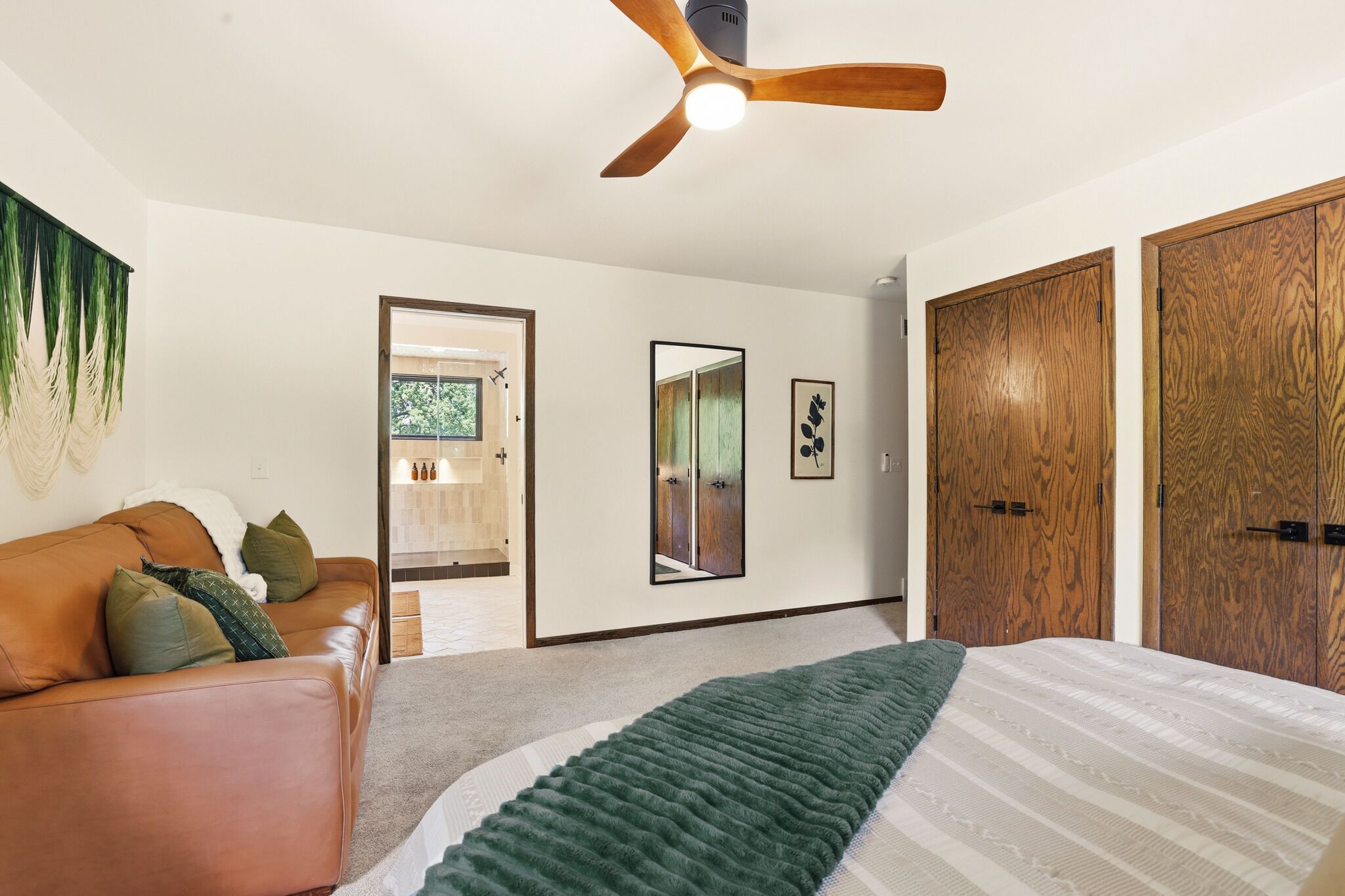 A bedroom with a bed, a ceiling fan, a wooden wardrobe, a mirror, and a door leading to a bathroom.