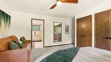 A bedroom with a bed, a ceiling fan, a wooden wardrobe, a mirror, and a door leading to a bathroom.