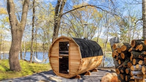 Eine Holzsauna mit Schornstein, eingebettet in eine bewaldete Landschaft mit einem See im Hintergrund.