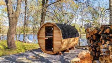 A wooden sauna structure with a chimney, set in a forested area with a lake in the background.