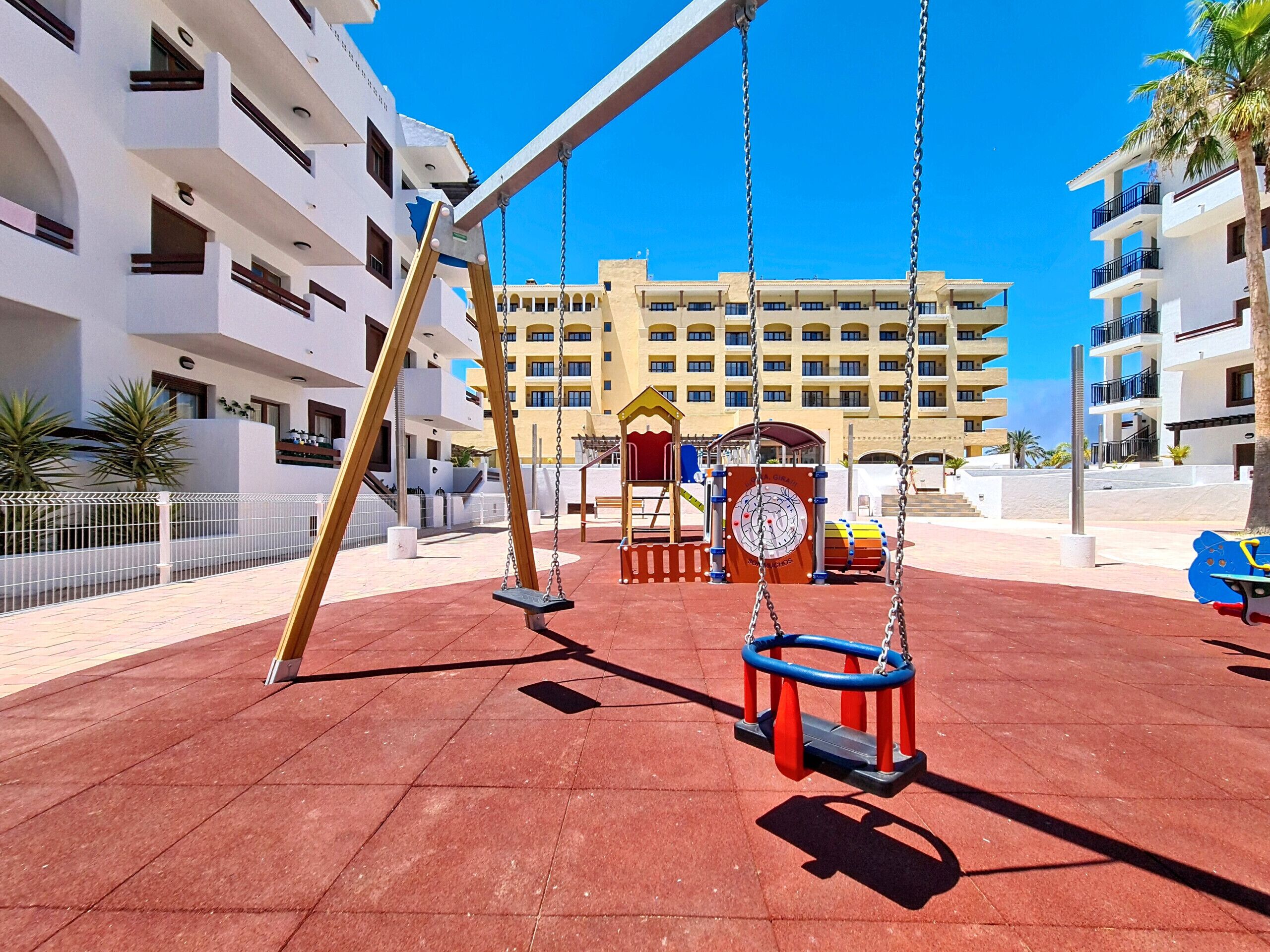 A playground with swings and a drum set, surrounded by multi-story buildings.