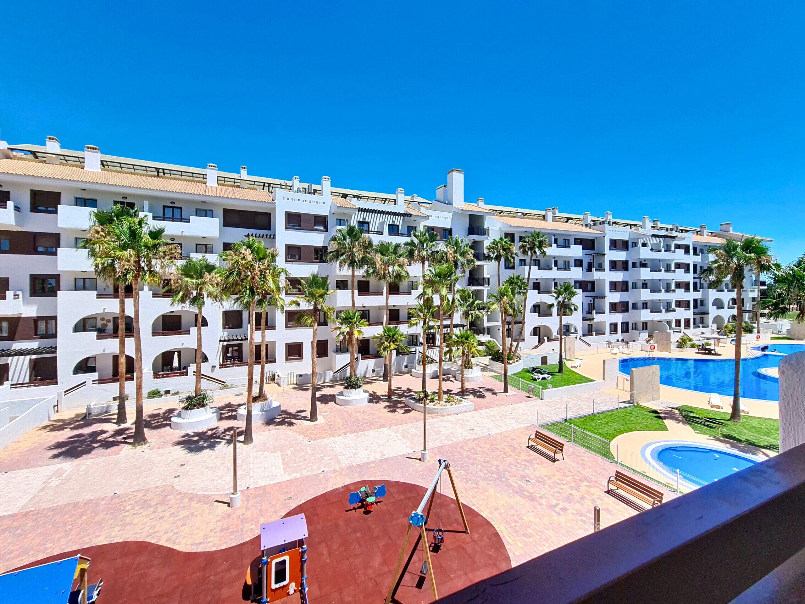 Un hôtel à étages, doté de plusieurs piscines extérieures, de palmiers et d’un espace de jeux pour enfants.