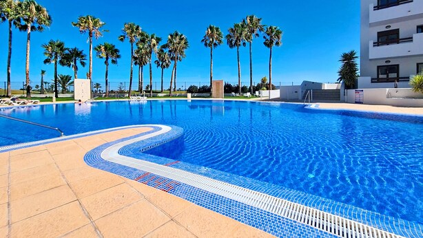 Una piscina con agua azul cristalina, rodeada de palmeras y un edificio blanco con balcones.