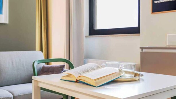 Ein Zimmer mit einer Couch, einem Tisch mit offenem Buch und einem Glas Wasser sowie einem Fenster mit Vorhängen.