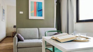 Ein Zimmer mit einer grauen Couch, einem kleinen Tisch mit einem Buch und Gläsern, einem gerahmten abstrakten Gemälde und einem Fenster mit Vorhängen.