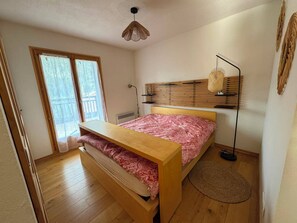 A bedroom with a wooden bed, a bedside table, a floor lamp, and a window with a view of a balcony. - New—Cozy Apartment in a Terrace Chalet in Les Houches—Mont Blanc (Les Houches)