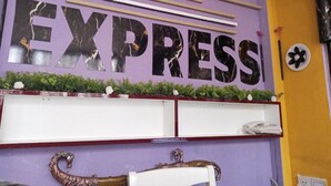 Un mur violet orné du mot « EXPRESS » en grandes lettres, une étagère blanche avec des objets décoratifs et une petite plante.