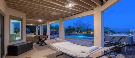A covered outdoor area with a hammock, seating, and a view of a swimming pool at dusk.