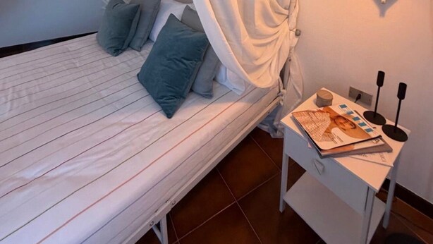 A bed with white curtains, a bedside table with a lamp, and a magazine. - A charming and modern apartment in the center of Teulada, just a few minutes from the beach (Teulada)