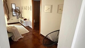 A room with a tiled floor, wooden ceiling beams, a bed with a white cover, a chair, and several framed pictures on the wall. - A charming and modern apartment in the center of Teulada, just a few minutes from the beach (Teulada)
