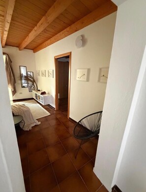 Ein Zimmer mit Fliesenboden, Holzbalkendecke, einem Bett mit weißer Bettdecke, einem Sessel und mehreren gerahmten Bildern an der Wand.