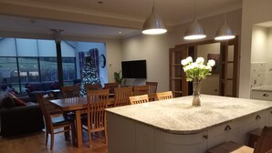 Una cocina moderna con una isla central, una mesa de comedor de madera y sillas, un televisor montado en la pared y un ventanal con vistas al exterior.