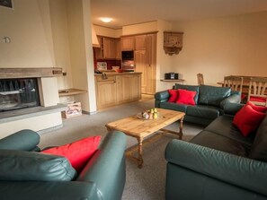 Ein Wohnzimmer mit Kamin, grünen Sofas mit roten Kissen und einem Holztisch. Im Hintergrund ist eine Küchenzeile zu sehen.