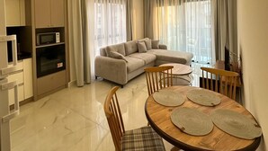 A modern living room with a sofa, dining table, and chairs, a kitchen area with appliances, and large windows with curtains. - "The Best Holiday Experience in Alanya & Mahmutlar." (Mahmutlar)