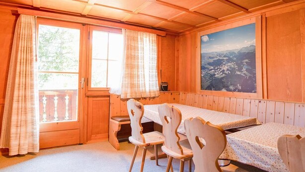 Ein Zimmer aus Holz mit einem Esstisch, Stühlen, einem Fenster und Blick auf eine Berglandschaft.