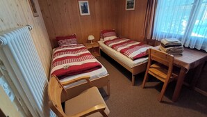 Ein Zimmer mit zwei Einzelbetten, einem Holztisch, einem Stuhl, einem Heizkörper und einem fenster mit Vorhängen.