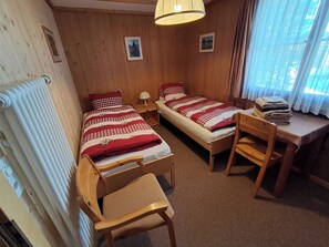 Ein Zimmer mit zwei Einzelbetten, einem Holztisch, einem Stuhl, einem Heizkörper und einem fenster mit Vorhängen.