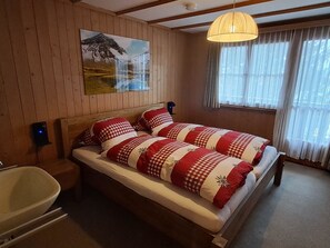 A double bed with checkered bedding, a wooden headboard, a mounted painting, a bedside lamp, a window with sheer curtains, and a wooden floor. - Chalet Suveran 2 Appartement/Fewo, Dusche oder Bad, WC, 6-Bettwohnung Suveran 2 by Interhome (Arosa)