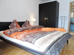 Ein Bett mit gemusterten Betttextilien, ein schwarzer Kleiderschrank und ein Nachttisch mit einer Lampe.