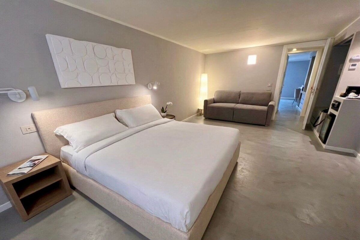 A modern hotel room with a large bed, a sofa, a small table, and a wall-mounted artwork.