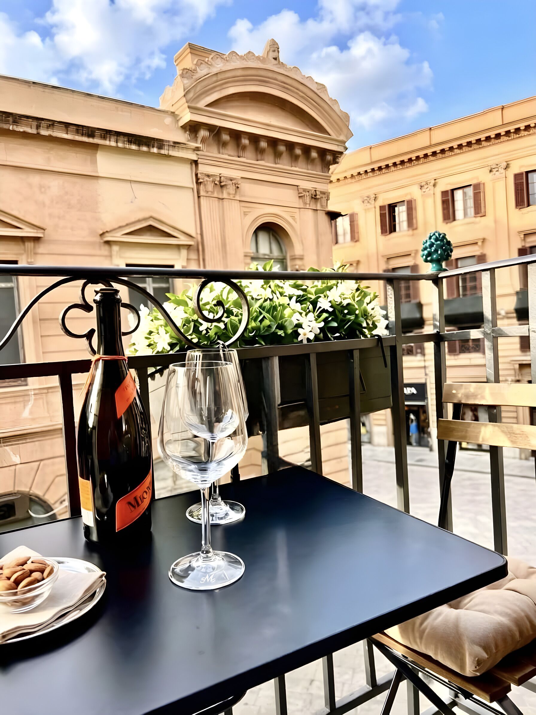 Ein Tisch mit einer Flasche und zwei Gläsern, eine Schale Mandeln sowie ein Kissen auf einem Balkon mit Blick auf Gebäude und Grünflächen.