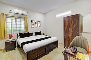 A hotel room with a bed, a wardrobe, a desk with a kettle, a chair, and a window with curtains.