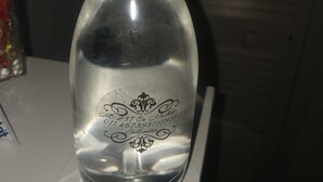 A clear glass bottle with a white cap, engraved with a logo and text, placed on a white surface.