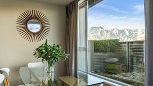 A modern room with a glass table, white chairs, a mirror with a sunburst frame, and a view of mountains through large windows. - Adelaide Courtyard | Walk to Town - Free Parking (Queenstown)