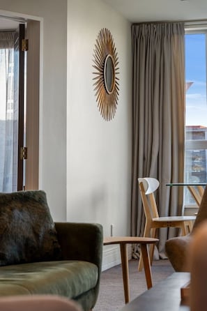 Ein modernes Wohnzimmer mit einer Couch, einem Esstisch mit Stühlen, einem dekorativen Spiegel und Blick auf die Stadt durch große Fenster.