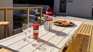 A table with a bottle of wine, glasses, a metal container with flowers, and a plate of food. - Rustic 3 Bedroom Beach Cottage, perfect for a peaceful getaway. (Birkenhead, Gansbaai)
