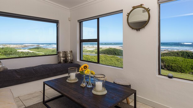 A room with a view of the ocean, a large window, a round mirror, a dark wooden table, and a cushioned bench. - Rustic 3 Bedroom Beach Cottage, perfect for a peaceful getaway. (Birkenhead, Gansbaai)