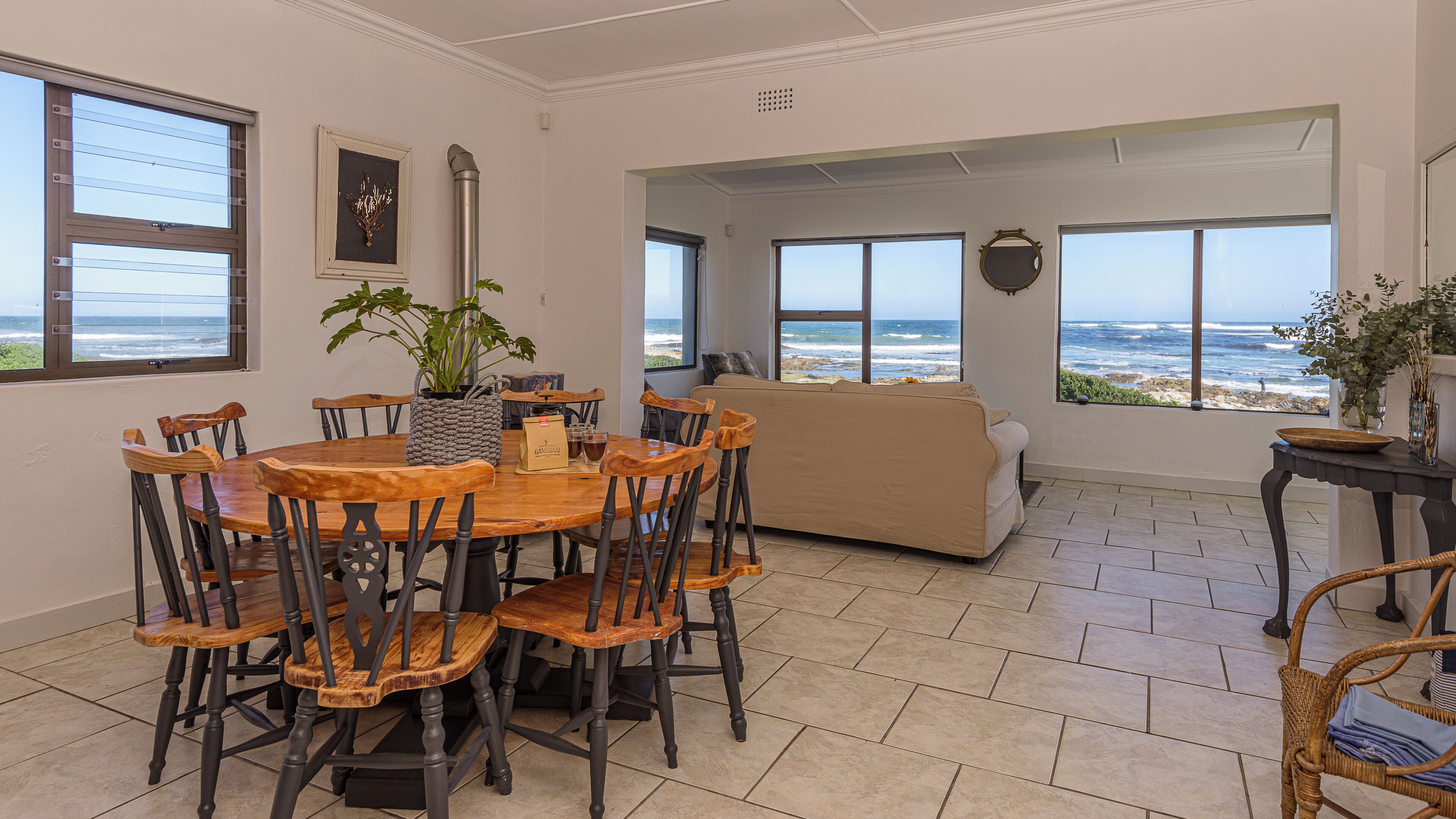 Un comedor con mesa y sillas de madera, un sofá color beige y ventanas amplias con vistas al océano.