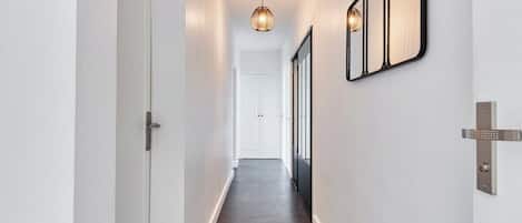 A hallway with a dark floor, white walls, a door on the left, and a wall-mounted light fixture on the right.